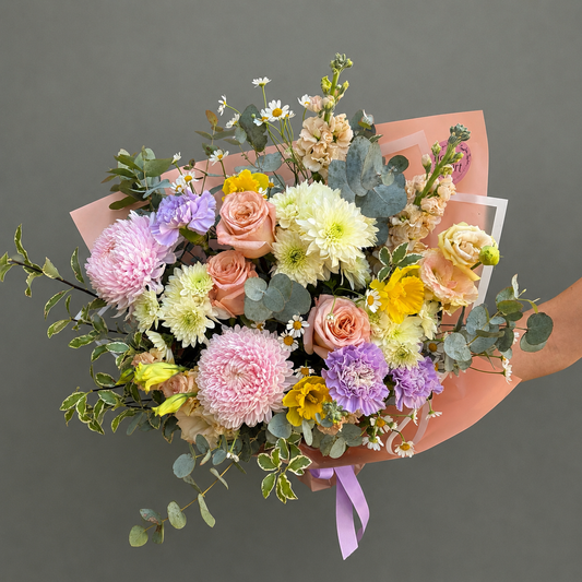 A vibrant floral bouquet with pink, mauve yellow, and apricot flowers, green eucalyptus leaves, and a lavender ribbon, wrapped in peach paper and held by a hand against a solid gray background.