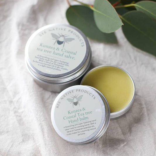 A small open tin container of hand salve with a beige color texture, placed on a linen surface with olive leaves in the background.