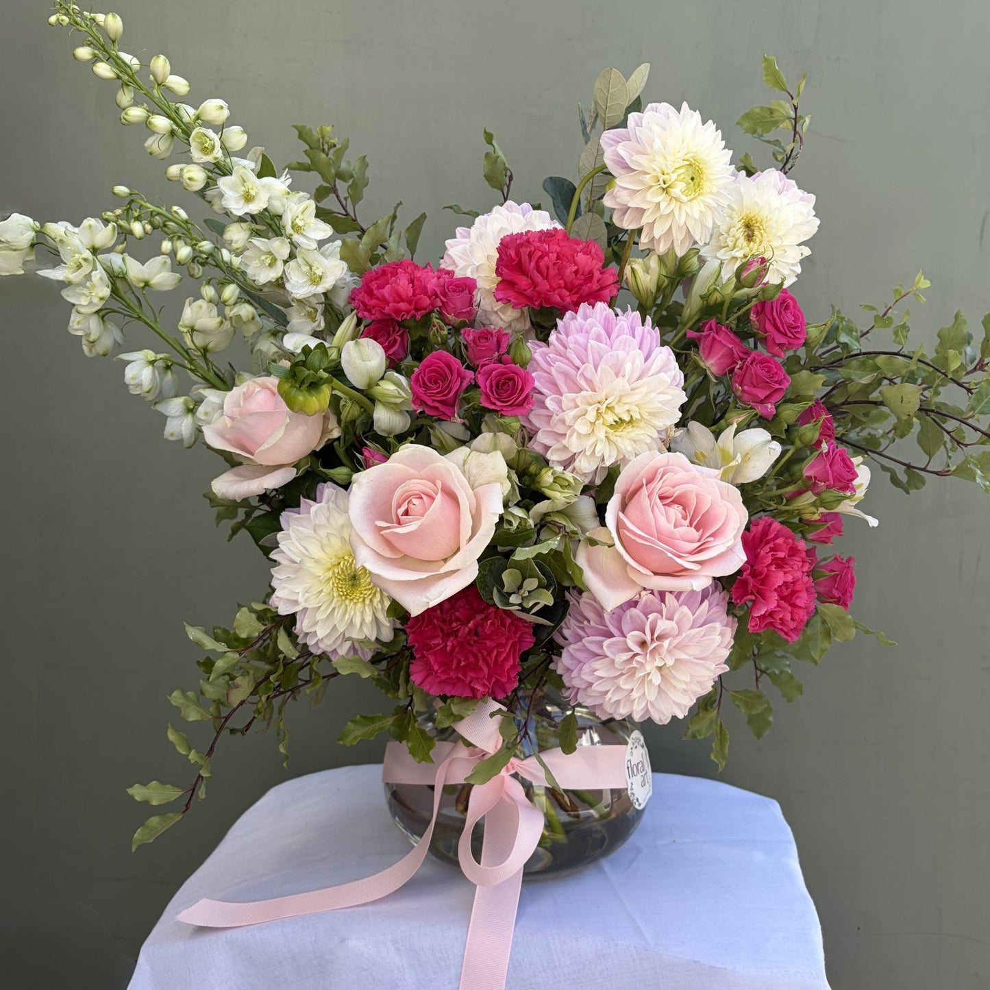Bouquet of pink and white flowers in a clear vase with a pink ribbon on a white surface against a green background