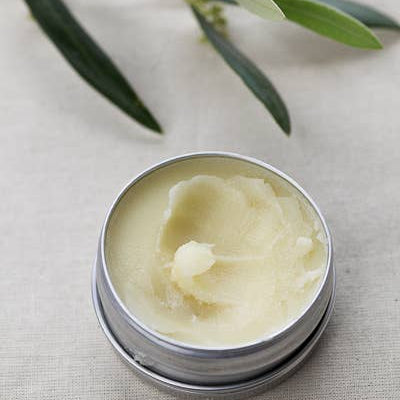 A small open tin container of hand salve with a beige color texture, placed on a linen surface with olive leaves in the background.