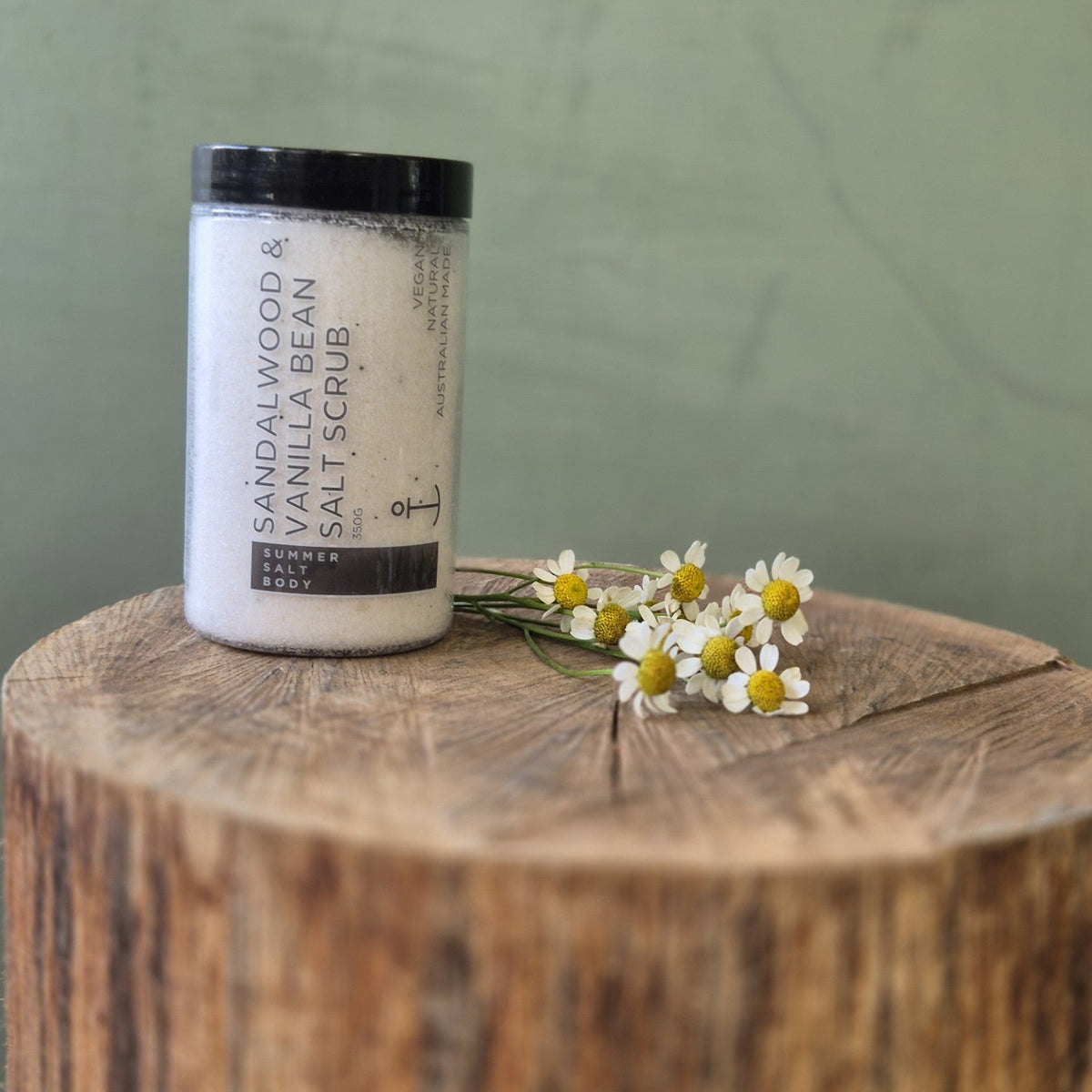 Jar of sandalwood and vanilla bean salt scrub on a wooden surface with flowers.