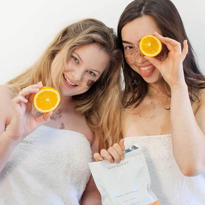 Two women wrapped in towels holding oranges and a package, smiling against a white background.