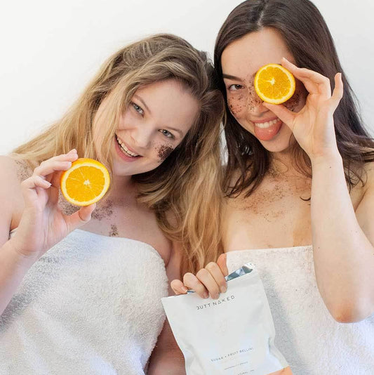 Two women wrapped in towels holding oranges and a package, smiling against a white background.