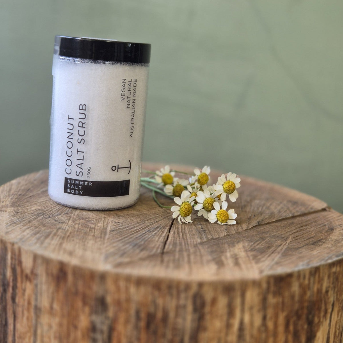 Coconut salt scrub jar with flowers on a wooden block against a green background