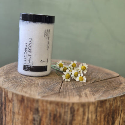 Coconut salt scrub jar with flowers on a wooden block against a green background
