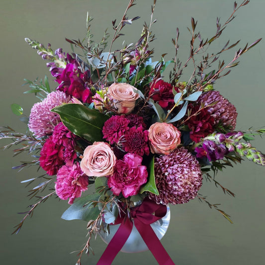 Bouquet of pink and purple flowers arrannged in a glass fishbowl vase with greenery on a plain background