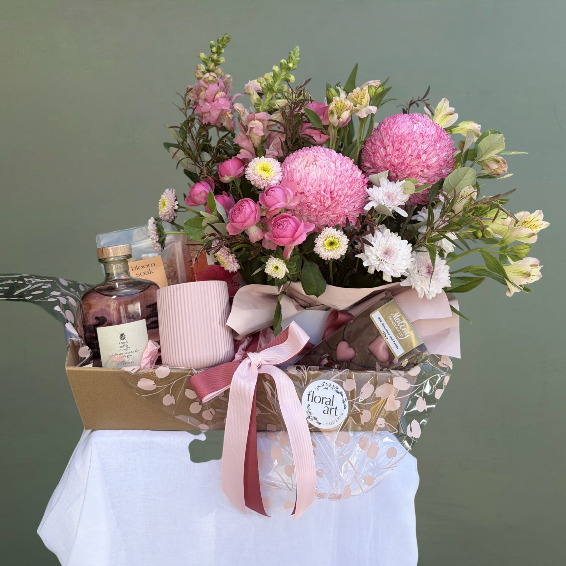 Gift basket with flowers, Pink Gin, Bath soak, candles, and chocolate on a white tablecloth against a green background