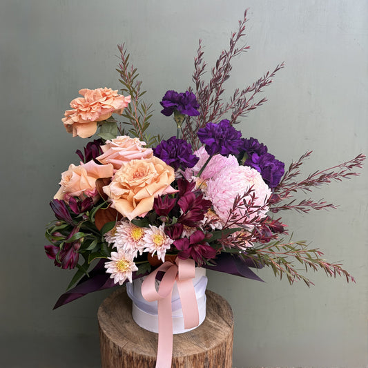  flowers arranged in hat box with peach, purple, and pink colors on a wooden stump against a green background
