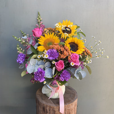 Colorful flower bouquet with sunflowers, roses, and hydrangeas on a wooden stump against a green background.