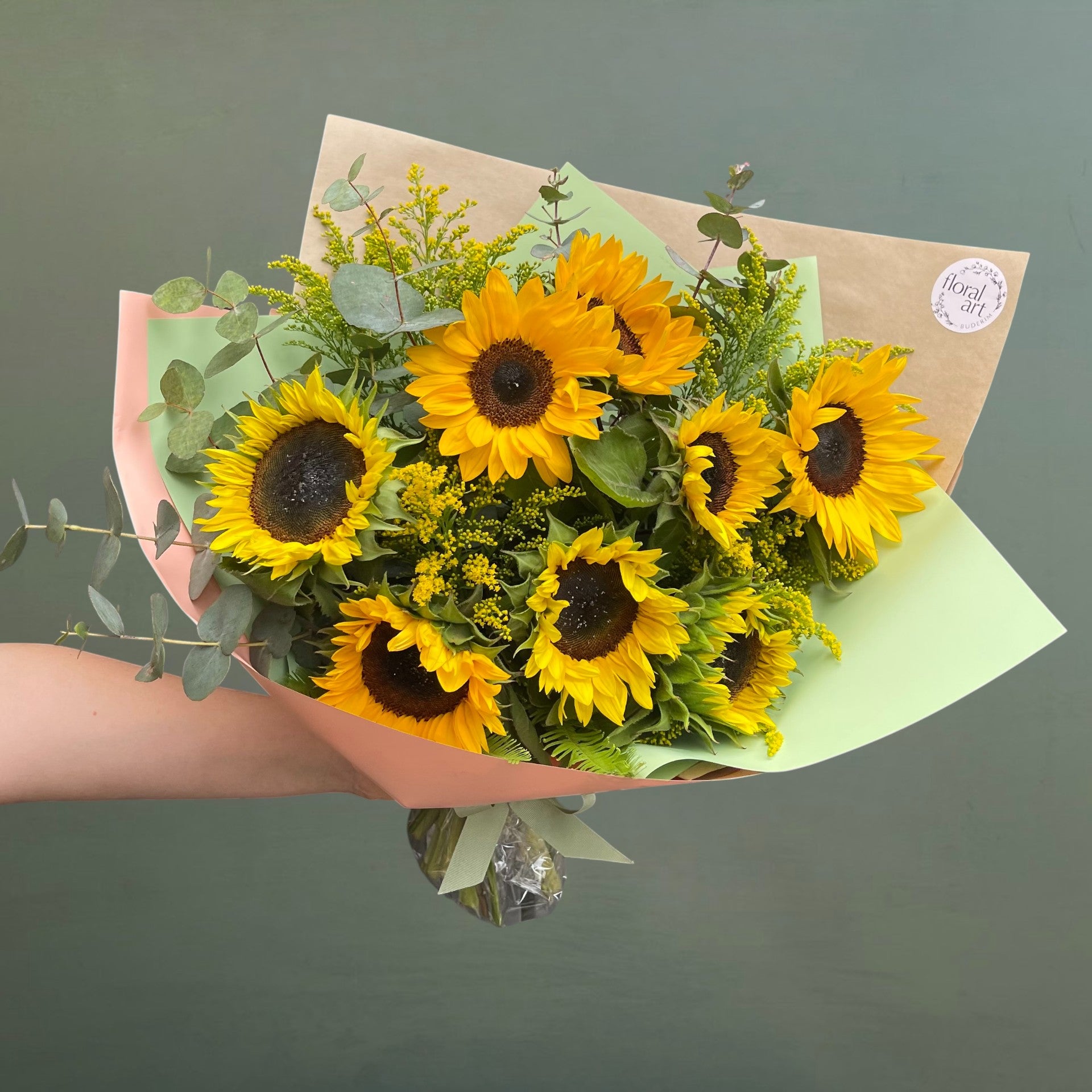 Bouquet of sunflowers wrapped in brown paper with a green leaf detail, held against a dark background