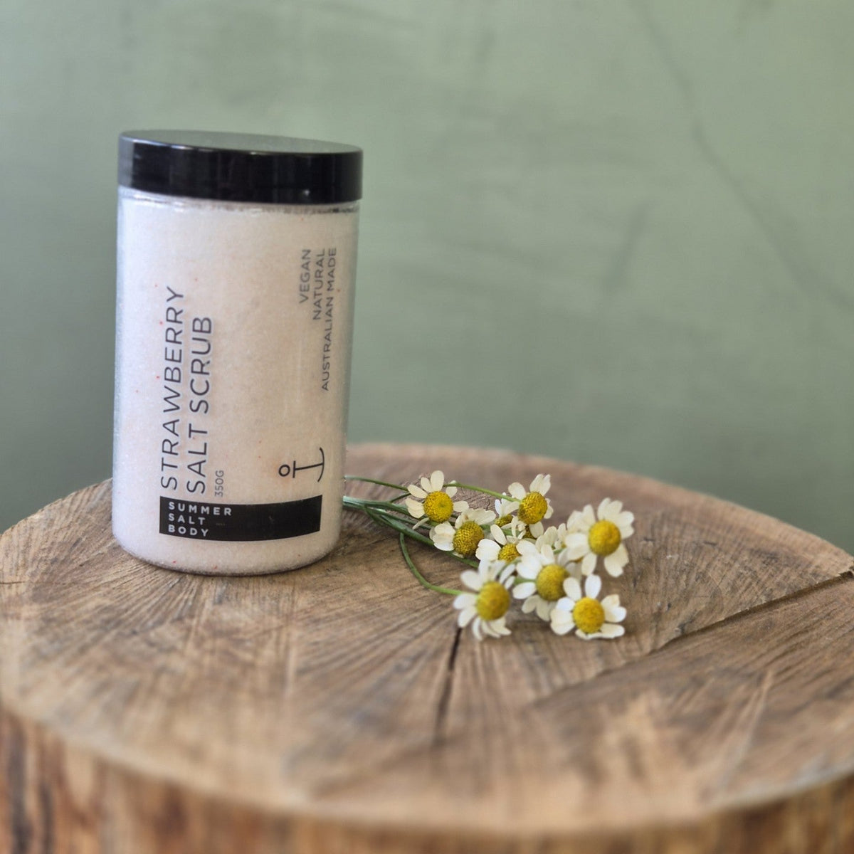 Strawberry salt scrub container on a wooden block with flowers against a green background