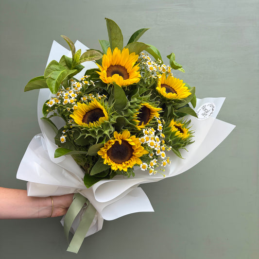 Bouquet of sunflowers with greenery and small white flowers, held against a plain background